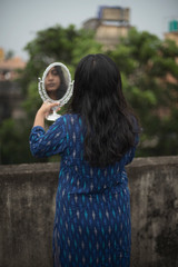 Indian woman looking her face on a handheld mirror standing on rooftop in front of a green tree wearing a blue dress in a cloudy afternoon. Indian lifestyle © abir