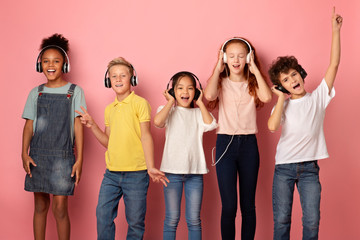 Multiethnic schoolchildren in headphones listening to music or audio books over pink background