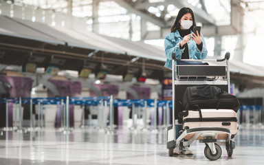 Asian tourist woman with mask using mobile phone standing alone with luggage trolley at the airport...