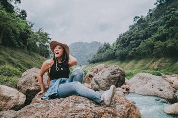 Naklejka na ściany i meble Happy Traveler Asian woman with backpack laughing while sitting on the rock by river in mountain during vacation. Travel in forest concept