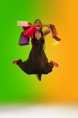 Jumping high, shocked with shopping bags. Portrait of young woman in neon light on gradient backgound. The human emotions, black friday, cyber monday, purchases, sales, finance concept. Trendy colors.