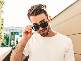 Portrait of handsome smiling stylish hipster lambersexual model.Man dressed in white T-shirt. Fashion male posing in the street in sunglasses 