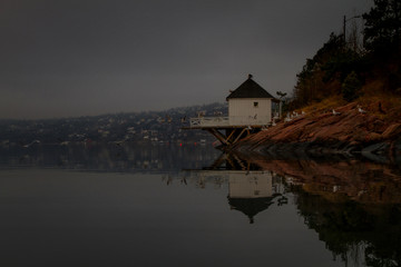 A white bathing house reflects in the calm surface of the fjord