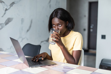 Beautiful young black woman using laptop computer on kitchen while drink water