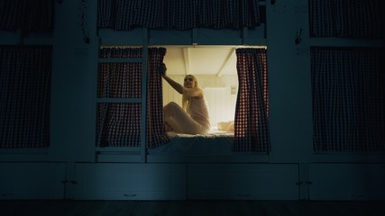 Young girl lays down in bed and closes the curtain in cozy hostel room