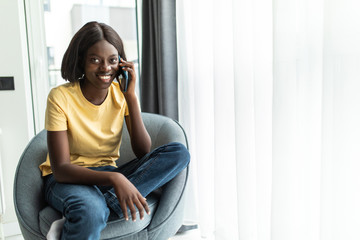 Obraz premium Relaxing weekend. Smiling african woman talking on mobile phone and enjoying coffee while resting in modern armchair at home