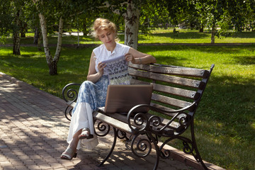 Woman sits on a park bench knitting wool clothes on a knitting needle and shows a knitted detail through a laptop. Distance learning