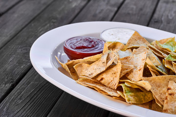 Triangular nachos chips with tomato ketchup and cheese sauce on a white plate and wooden background