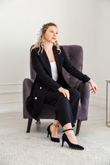 A blonde girl in a black suit and high-heeled shoes sits in a chair and poses at the camera