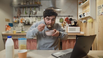 Young man working laptop computer and eats Corn Flakes Cereal at home kitchen.