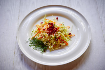 Fresh cabbage salad made from shredded white cabbage, carrots and berries on a white plate. Top view, wooden background, selective focus