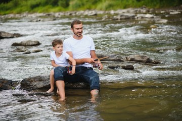 A father teaching his son how to fish on a river outside in summer sunshine. father's day.