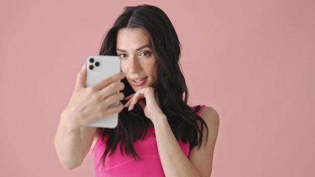 A cheerful young woman is taking selfie photo using her smartphone standing isolated over a pink background