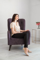 A beautiful brown-haired woman in dark trousers and a light blouse sits in an armchair and looks out the window.