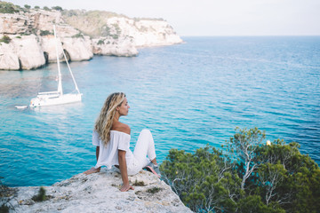 Carefree tourist spending day for enjoying scenery landscape views of Menorca recreating during adventure journey, Caucasian female solo traveller resting at rocky abyss near berth with yacht