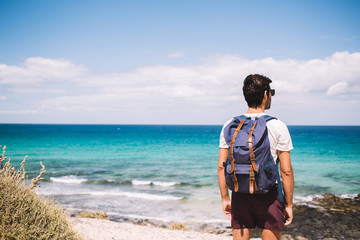 Back view of male wanderer with touristic rucksack standing at wild shoreline and admire picturesque scenery views of turquoise seascape, traveller with backpack looking at Mediterranean sea