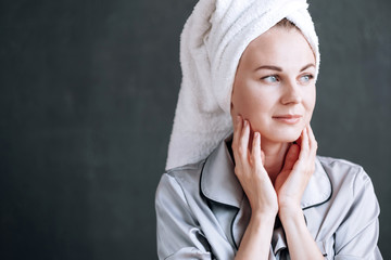 young woman 30 years old with a white towel on her head and gray pajamas on a dark background. touches the skin of the face with your fingers. healthy and clean skin concept