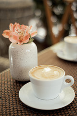 Morning cup of coffee latte and tender flowers in a summer cafe with bokeh in the background.