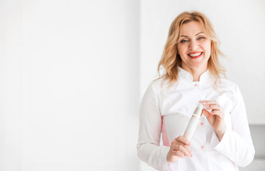 Stylish and beautiful woman beautician in a white medical suit smiles and looks into the camera. Woman holding a cosmetic product in her hands