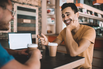 Happy cheerful caucasian man laughing during conversation with friend in coffee shop. colleagues students talking to each other enjoying prosperous startup project using technology with blank screen.