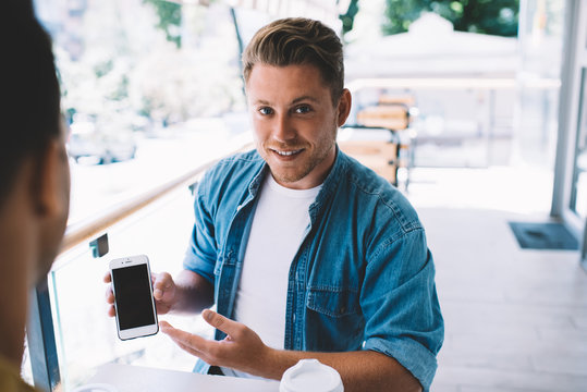 Portrait of cheerful millennial hipster guy holding smartphone with mock up screen showing video to his friend on meeting, cropped image of men colleagues sitting together male holding mobile phone.
