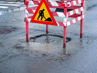Sunken sewage cover on asphalt road, wood frame around it with working ahead road sign.