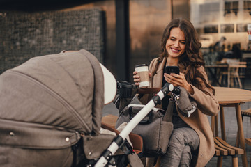 Young lovely mother with a stroller drinks coffee and talking on phone sitting in street cafe.