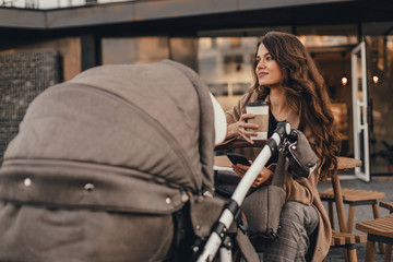 Young lovely mother with a stroller drinks coffee sitting in street cafe.