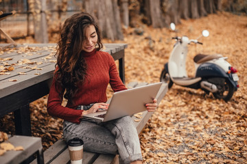Beautiful woman in cozy outfit works at laptop while sitting on chair near cafe in autumn park.