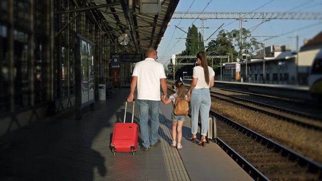 Family with a girl 5-6 years old and suitcases is walking along the platform. There are medical masks on the faces of passengers. Back view. Travel during the COVID-19 epidemic.