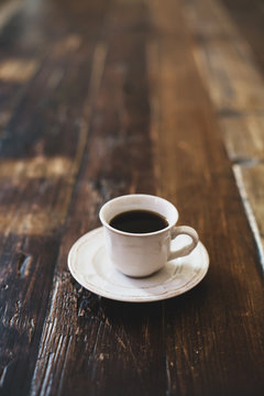 A white cup with black coffee and a vintage saucer are on a wooden brown table. Atmospheric image of coffee in warm colors. Copy space. Vertical image.