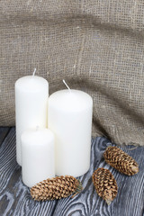 Three white candles stand on painted pine planks. Spruce cones are spread out nearby.
