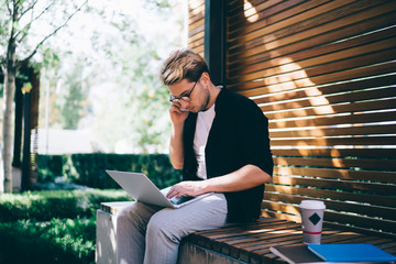 Young freelancer using laptop and talking on smartphone on bench