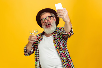 Time to drink and selfie. Portrait of senior hipster man in eyewear isolated on yellow studio...