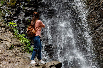 Woman is filming a waterfall on the phone with a backpack behind her back.