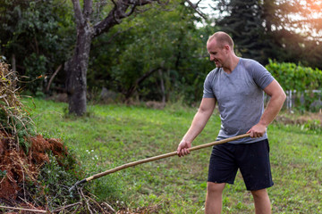 Photo of man gardener raking leaves from maple tree.Young man homeowner in garden backyard collecting of dry branches standing with rake on sunny day.Shot of man working with gardening rake in garden.