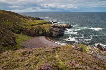 Fototapeta na wymiar Lonely Scottish beach near Gairloch with heather and grassy path in foreground, pink shingle beach and cliffs, Isle of Skye in far distance.