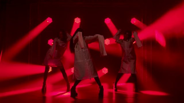 Silhouettes of a three charming womans in white shirt are dancing on the stage. Shot in a dark studio with smoke and neon lighting. Red neon light effects. Dance show.