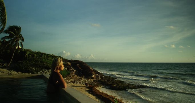 Woman jumps up to lean against the edge of a swimming pool that sits overlooking ocean and beautiful sun set