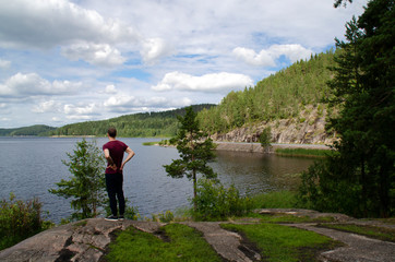 A young man stands admiring the beautiful views. Lake, fir trees on the hills, blue sky with clouds. Northern landscapes in Karelia, Russia.