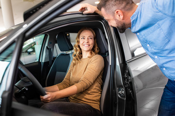 Smiling mid-adult couple buying a new car from dealership, woman sitting inside the vehicle while...