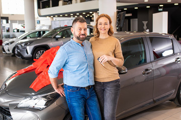 Portrait of smiling adult couple standing in front of a vehicle after buying a new car at dealership.