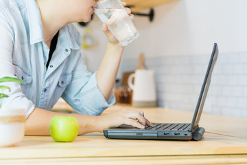 Latin woman drinking water while using her laptop, a fruit on the table.