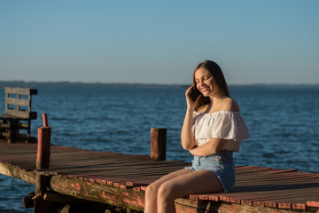 young blonde woman sitting on a pier using her cell phone
