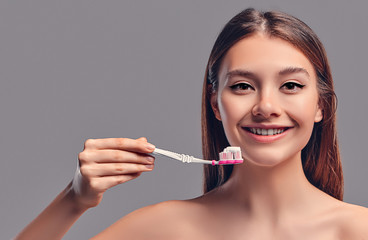 Young attractive brunette girl with loose hair uses a toothbrush and paste isolated on a gray background.