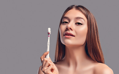 Obraz premium Young attractive brunette girl with loose hair uses a toothbrush and paste isolated on a gray background.