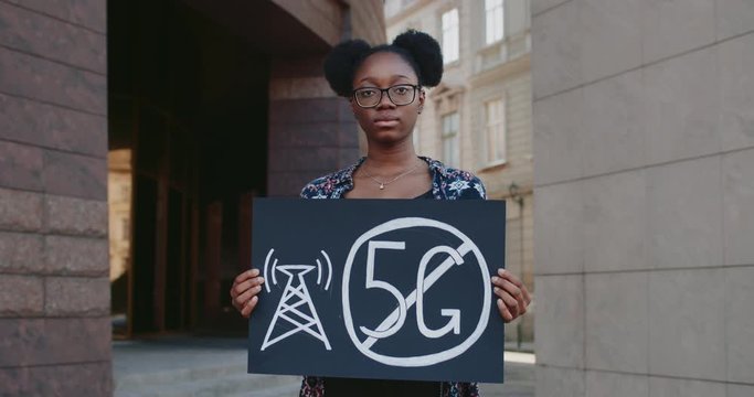 Portrait of millennial afro american woman holding placard with no 5g sign. Girl in glasses protesting against new technology and compatible antenna deployment while standing at street