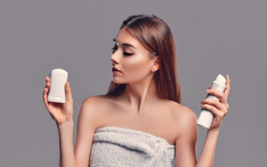 Young attractive brunette girl with loose hair with deodorant stick and antiperspirant aerosol isolated on gray background. Body care.