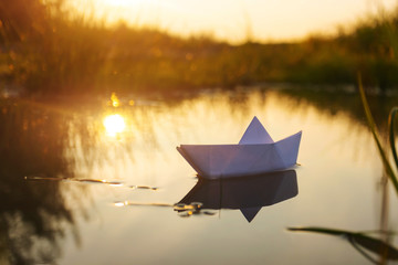 A paper boat floats on the water at dawn. Creative concept of outdoor recreation, river, travel. Cozy cute image in warm colors. Copspace.
