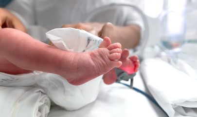 Nurse in blue gloves takes action to monitor and care for premature baby, selective focus on his feet. Newborn is placed in the incubator. Neonatal intensive care unit